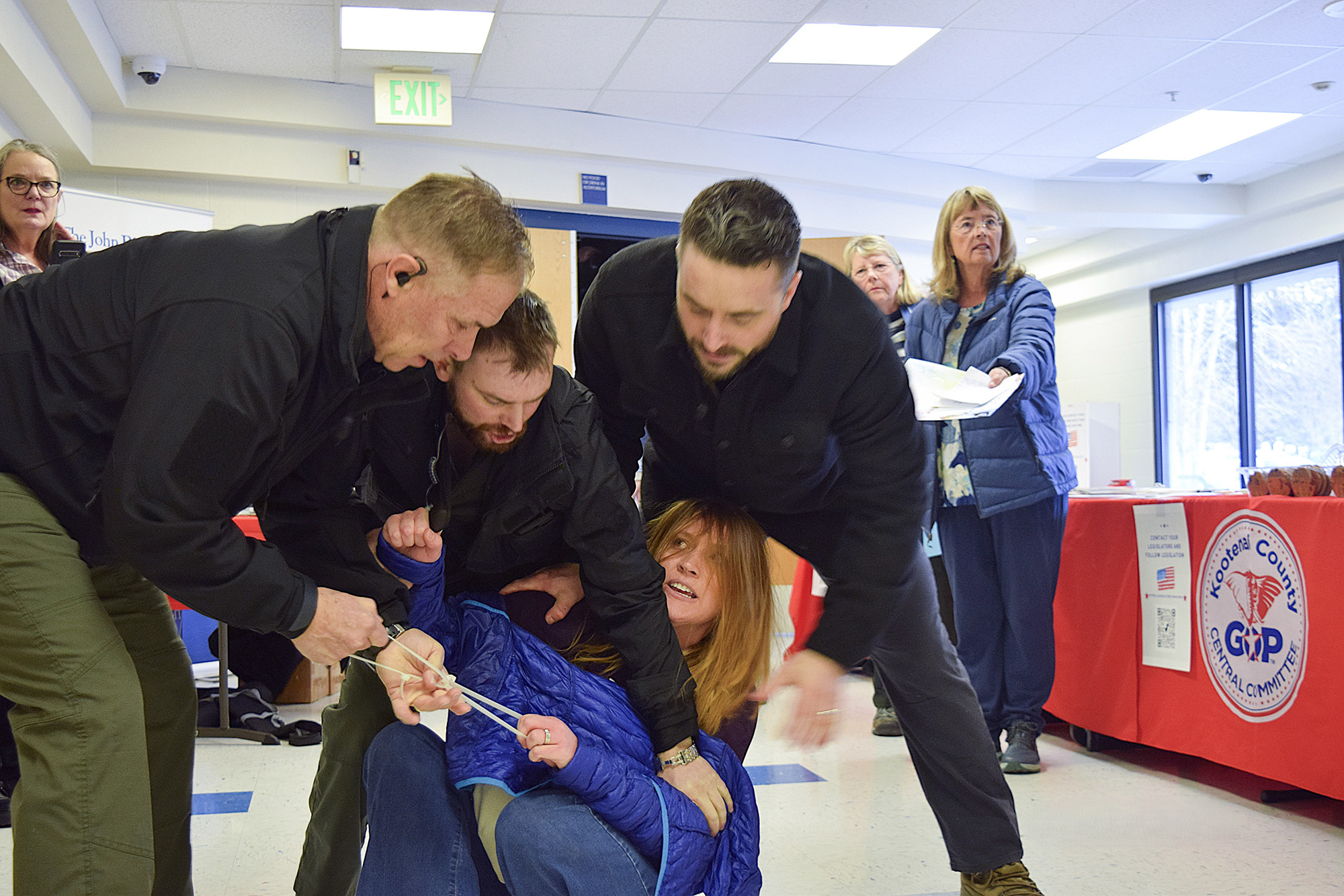 A woman is being grappled and dragged down a hall by three men, as people look on.