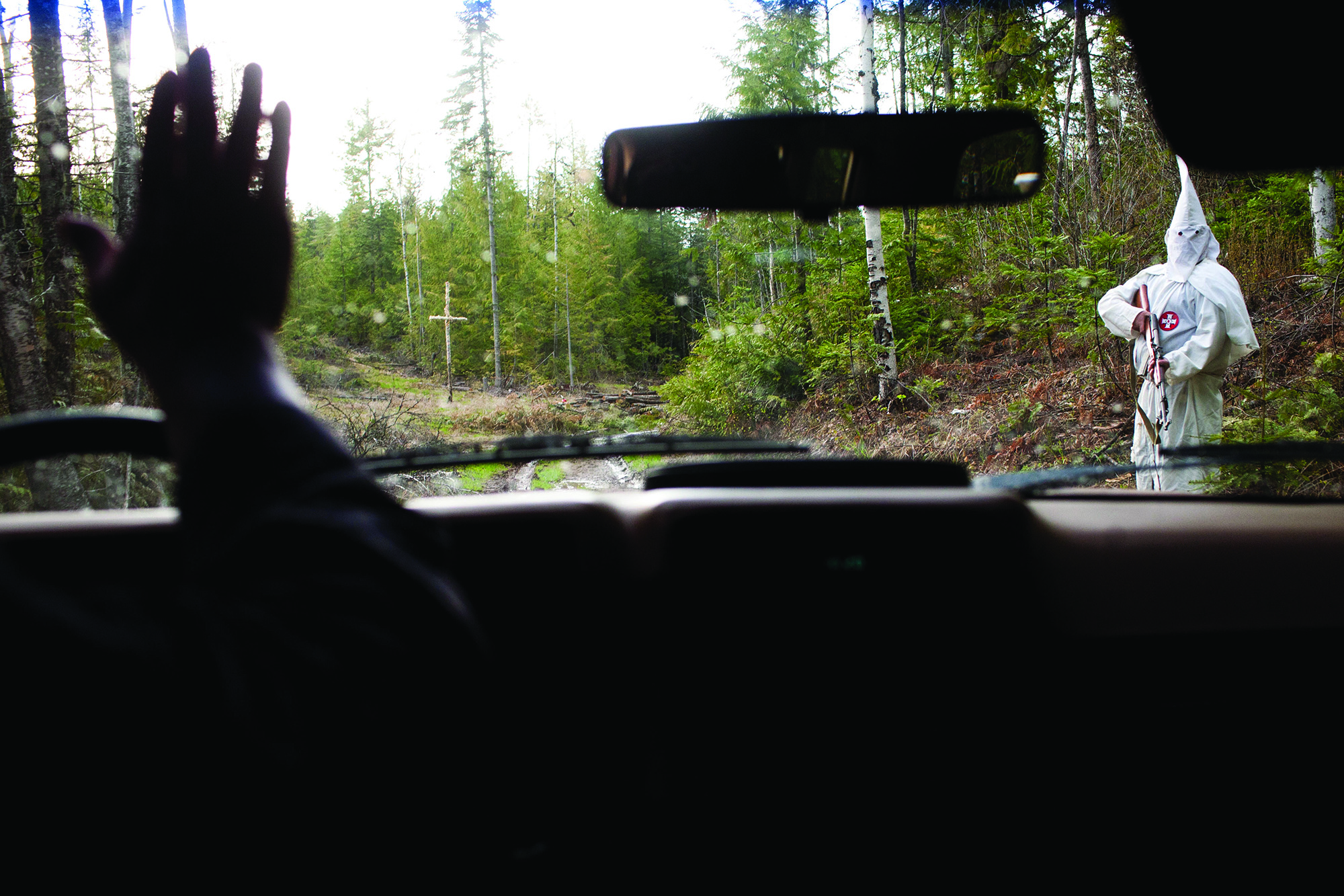 Silhouette of a man's hand from inside a car waving to a hooded KKK member bolding a firearm.