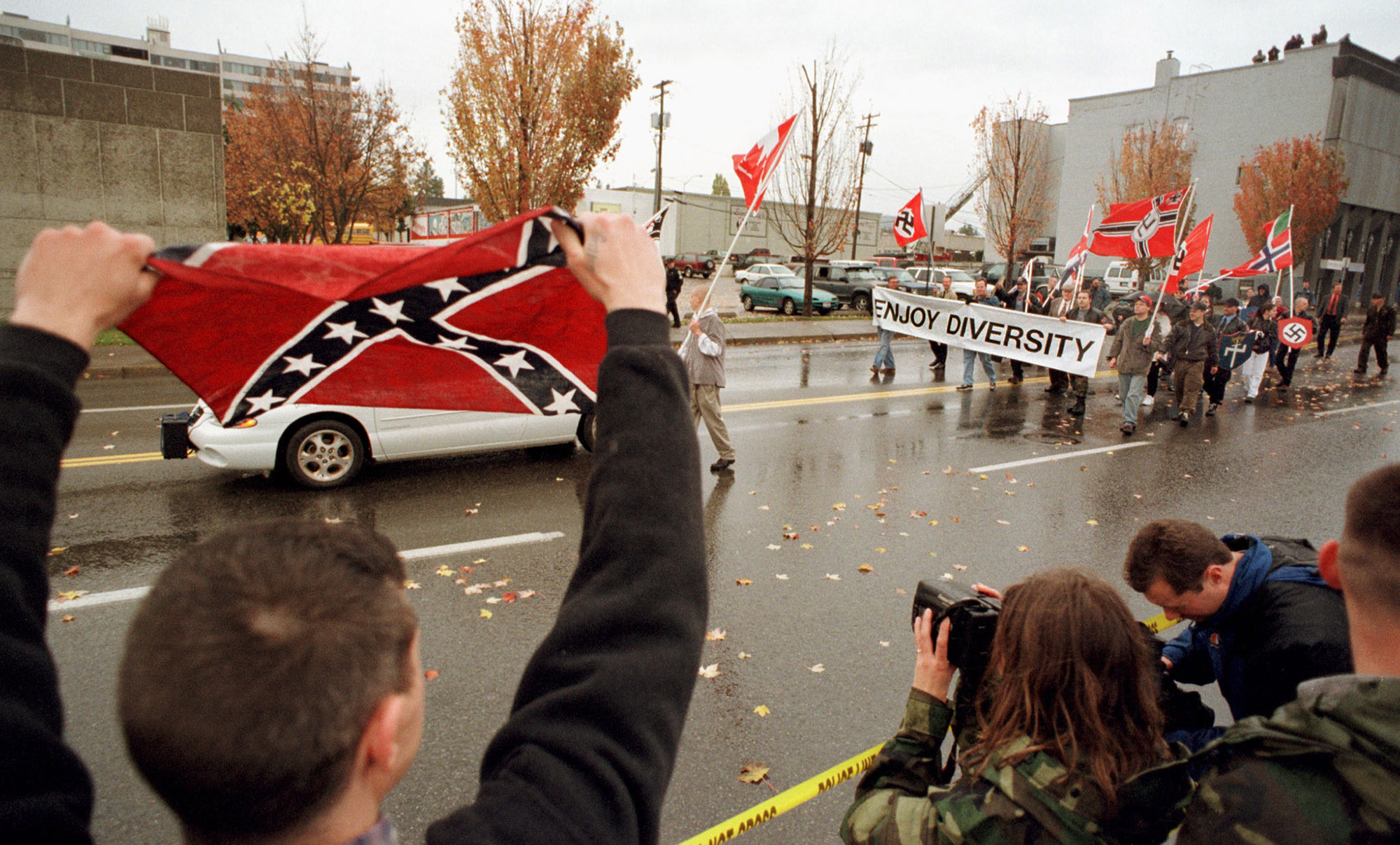 A crowd watches a parade of white nationalists holding signs and carrying Nazi flags on a downtown street; a man in the foreground raises a Confederate flag in support.