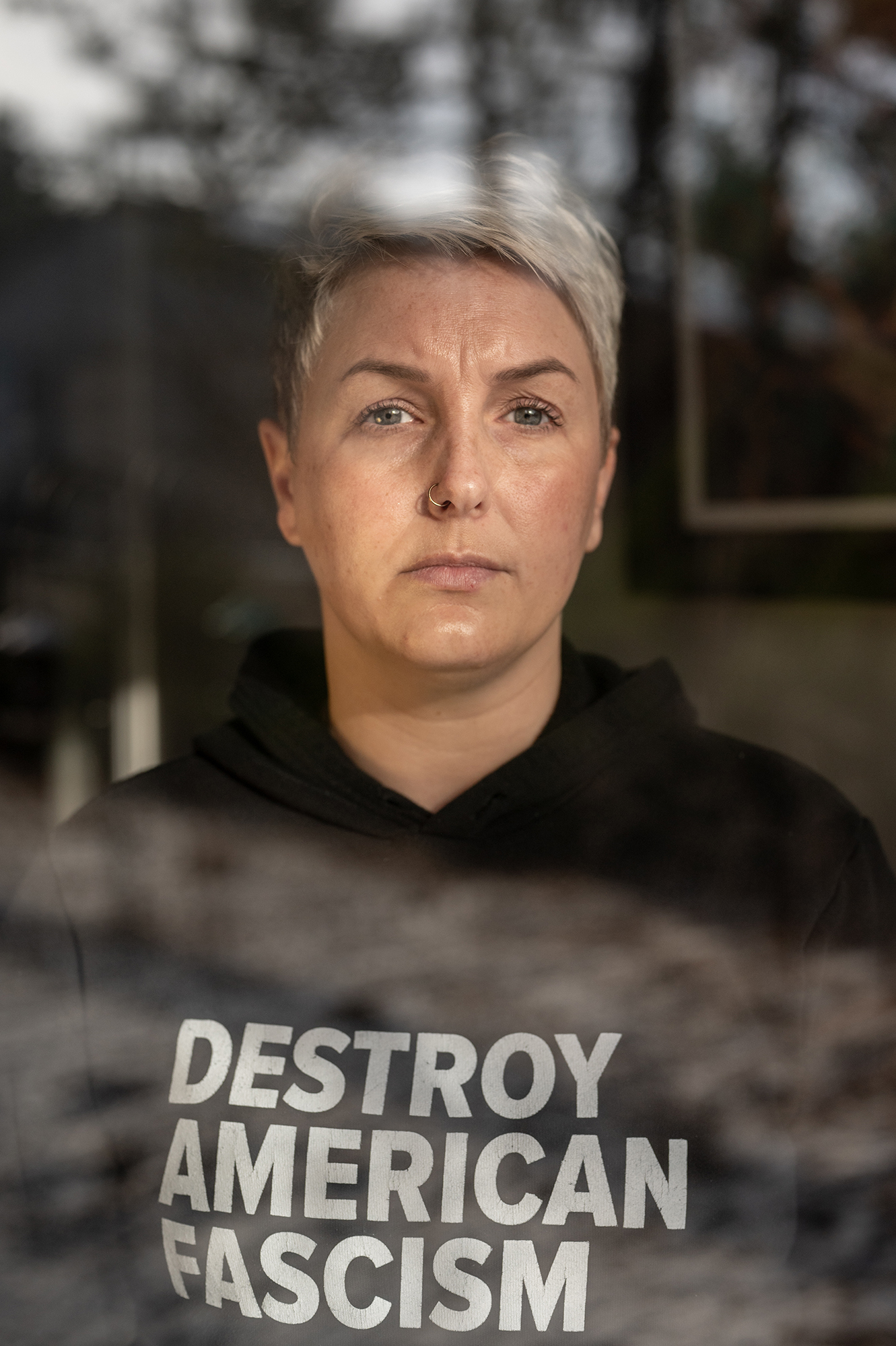 Portrait of a woman with short hair wearing a hoodie that reads DESTROY AMERICAN FASCISM through a window.