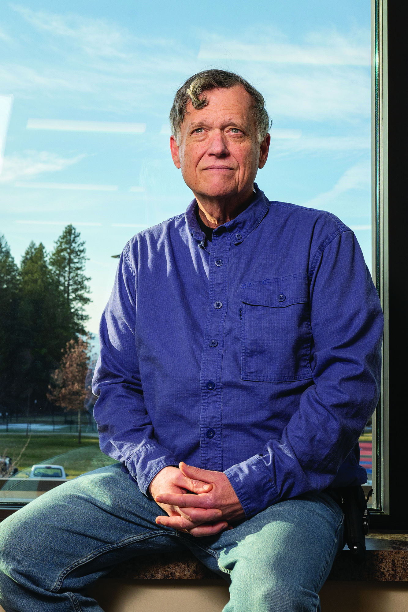 A middle aged man, with a serious expression, on jeans and a button down, sits on the ledge of a window.