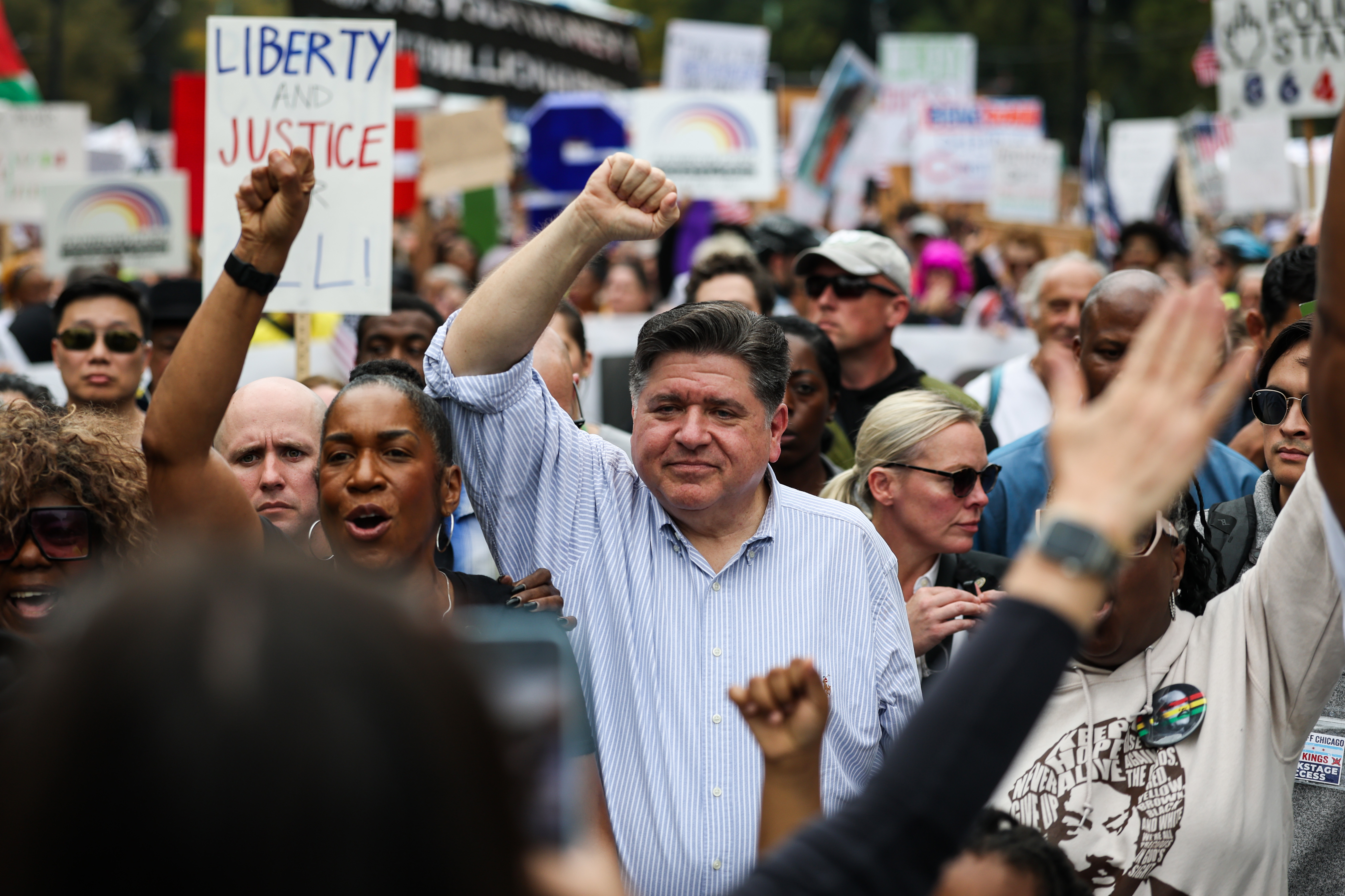 A middle-aged man wears a blue-striped button-down shirt with the sleeves rolled up. With his right fist raised, he is surrounded by a crowd of people of varying races, ages, and genders, some holding signs that have slogans such as "Liberty and Justice for All."