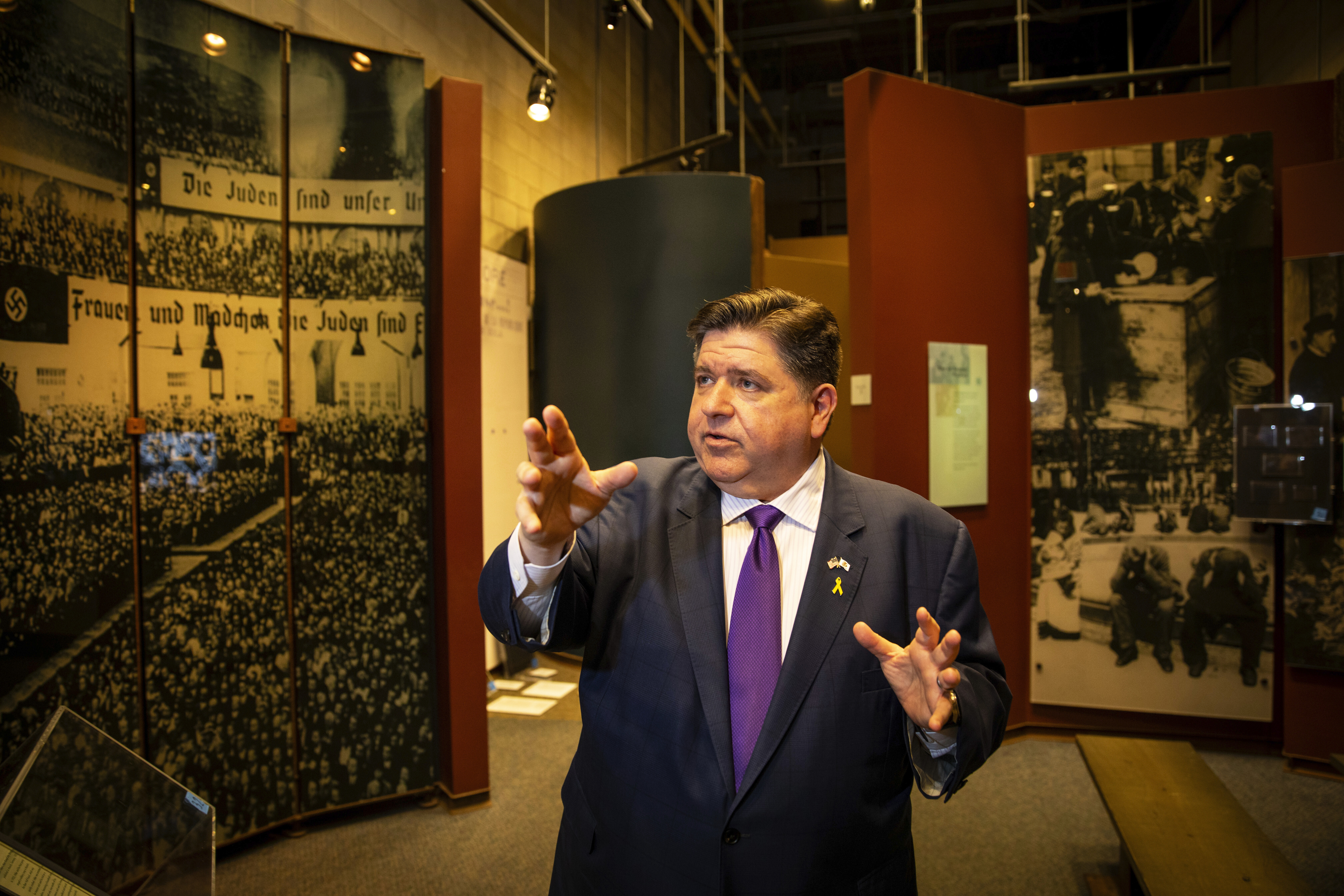 A middle-aged man wearing a dark suit and purple tie speaks in front of photos and displays nearly the height of the wall of Nazi rallies and oppression.
