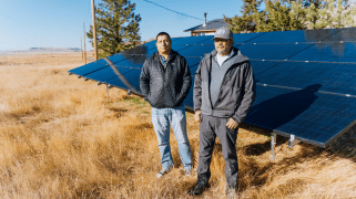 Two men stand in a field of brown grass in front of solar panels.