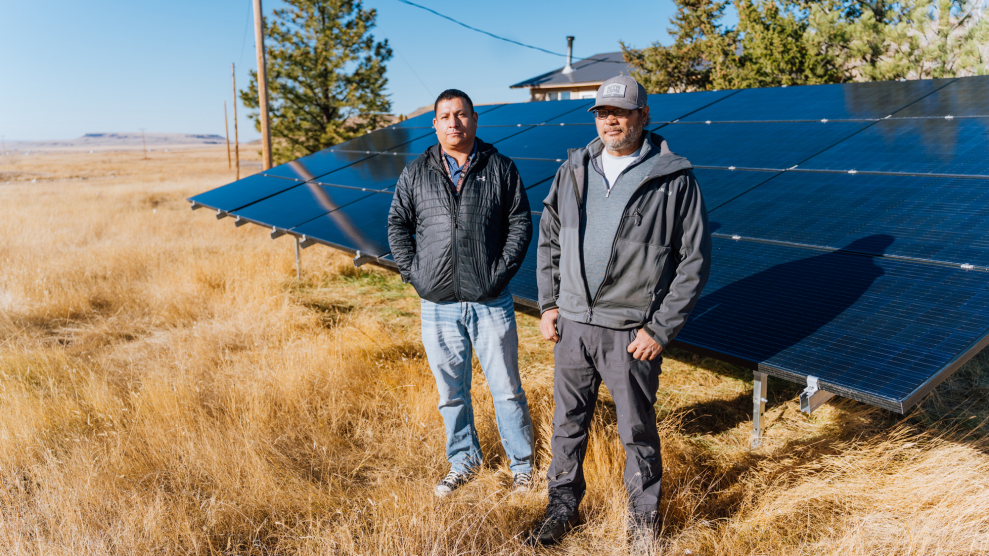 Two men stand in a field of brown grass in front of solar panels.