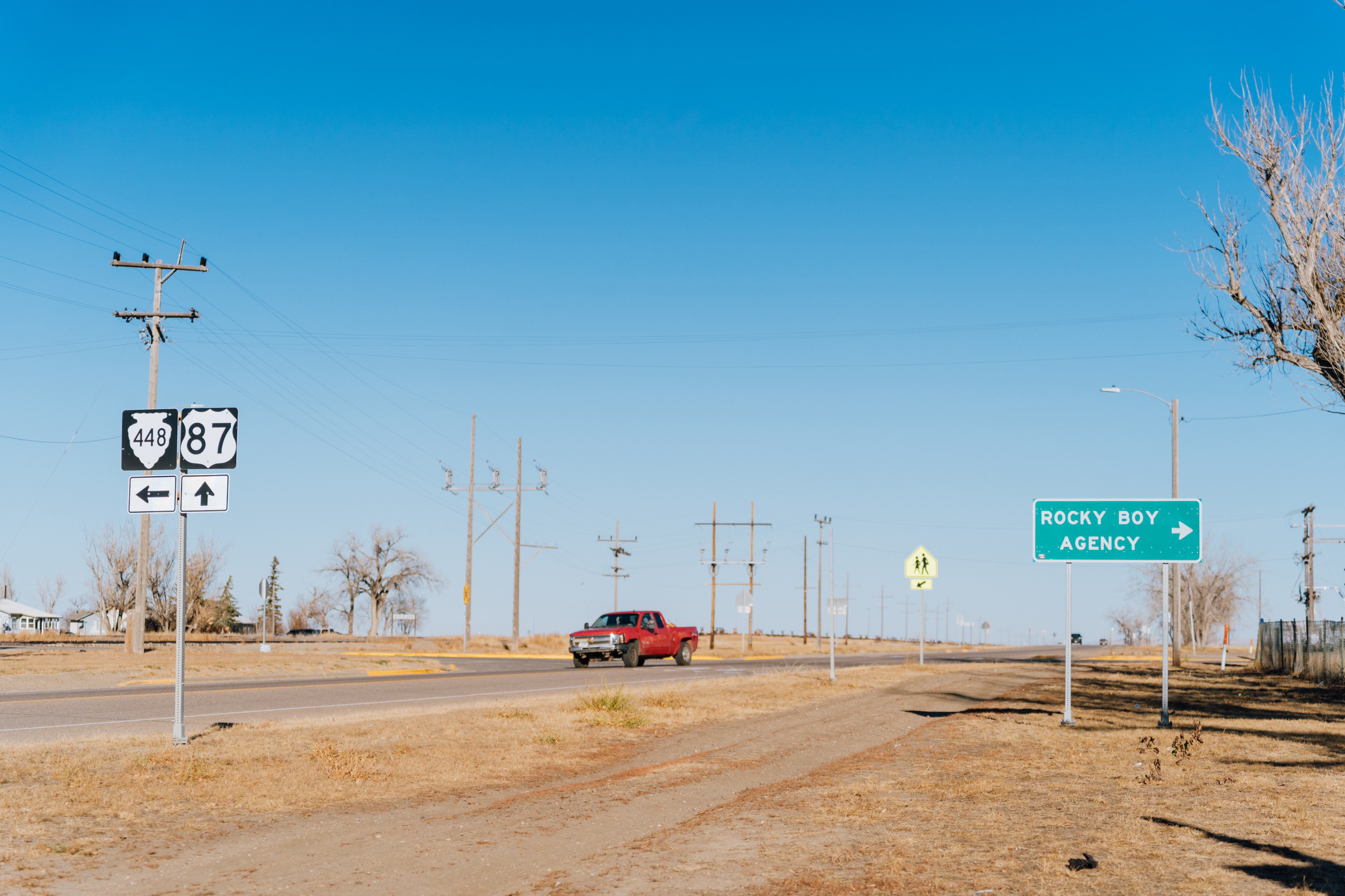 A red pickup truck drives down a desolate highway on a sunny day. A road sign pointing to the right reads, "Rocky Boy Agency."