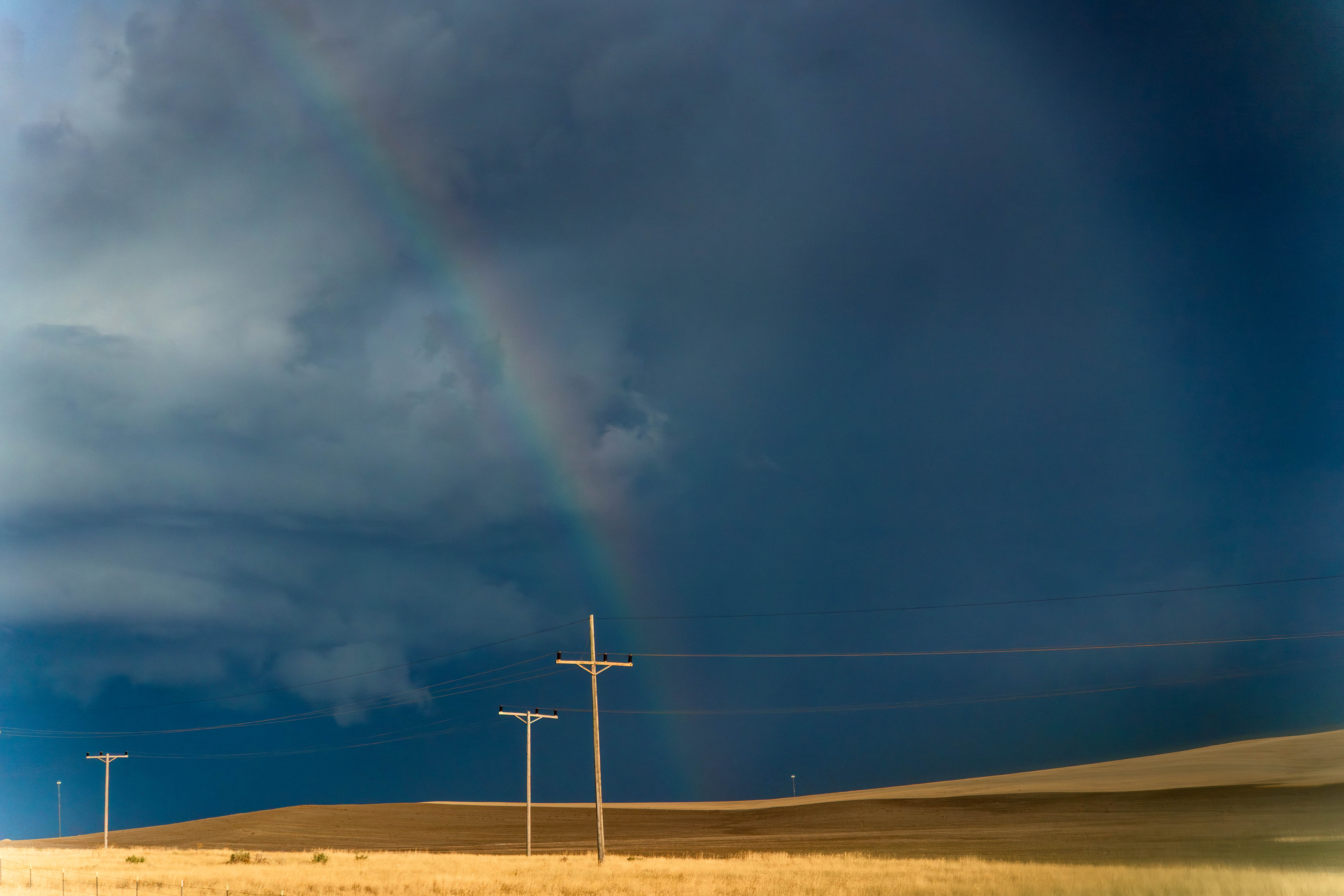 A faint rainbow and dark clouds hang over a golden field. Three power-line poles stand in the distance.