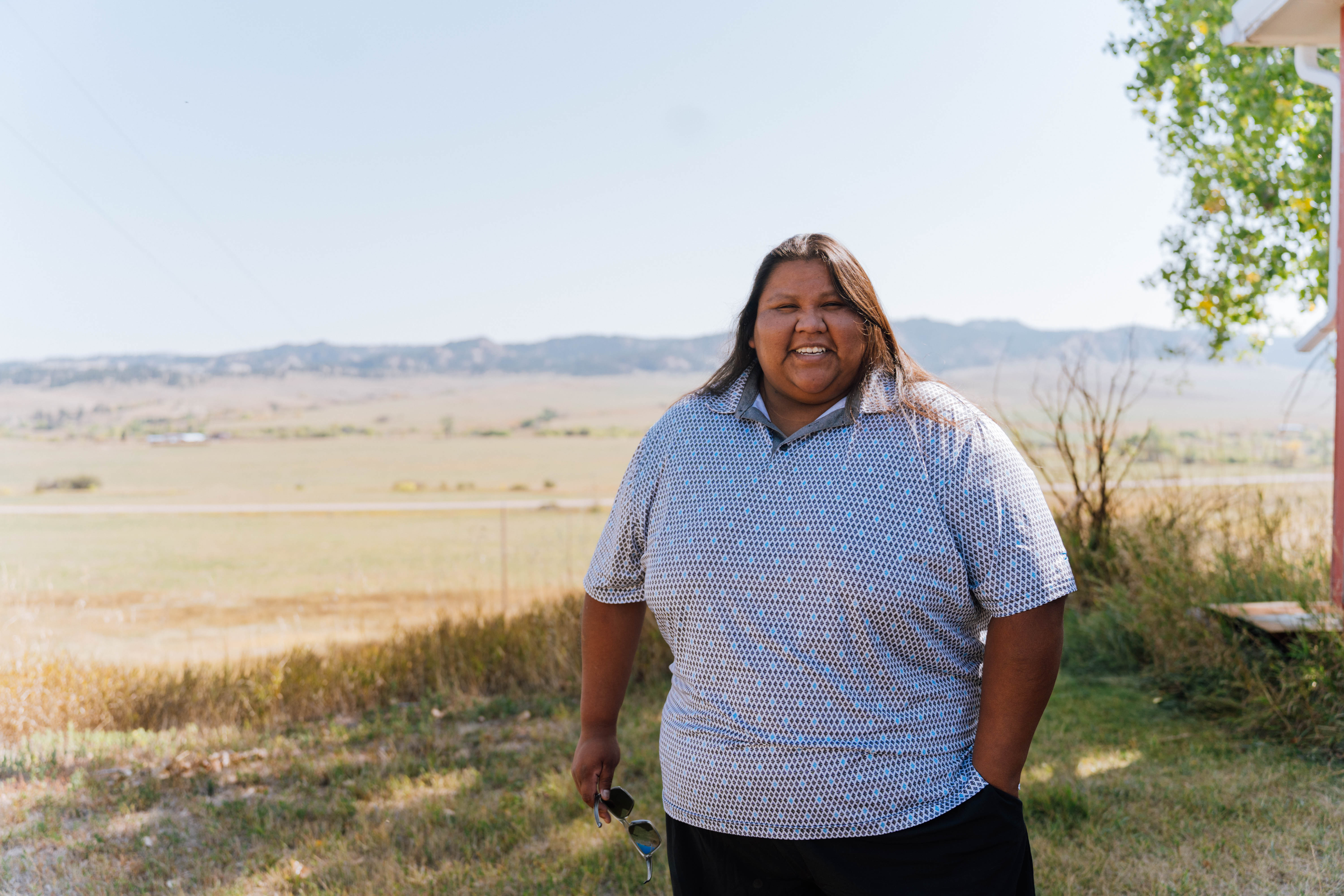 A woman smiles for a portrait, standing outside on a sunny day. A valley of green grass and distant hills are visible behind her.