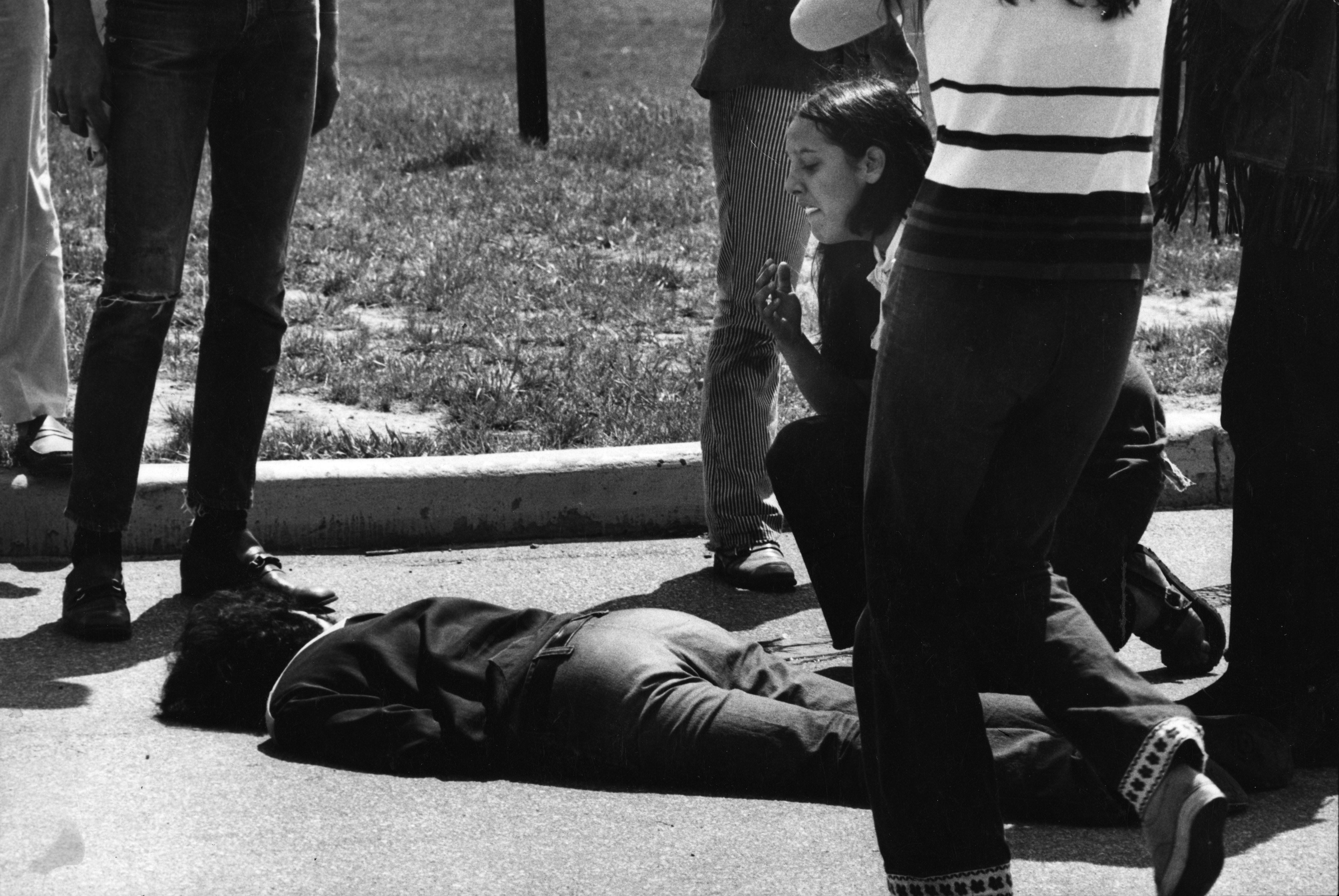 Woman kneels over the body of a person shot, laying on the ground.