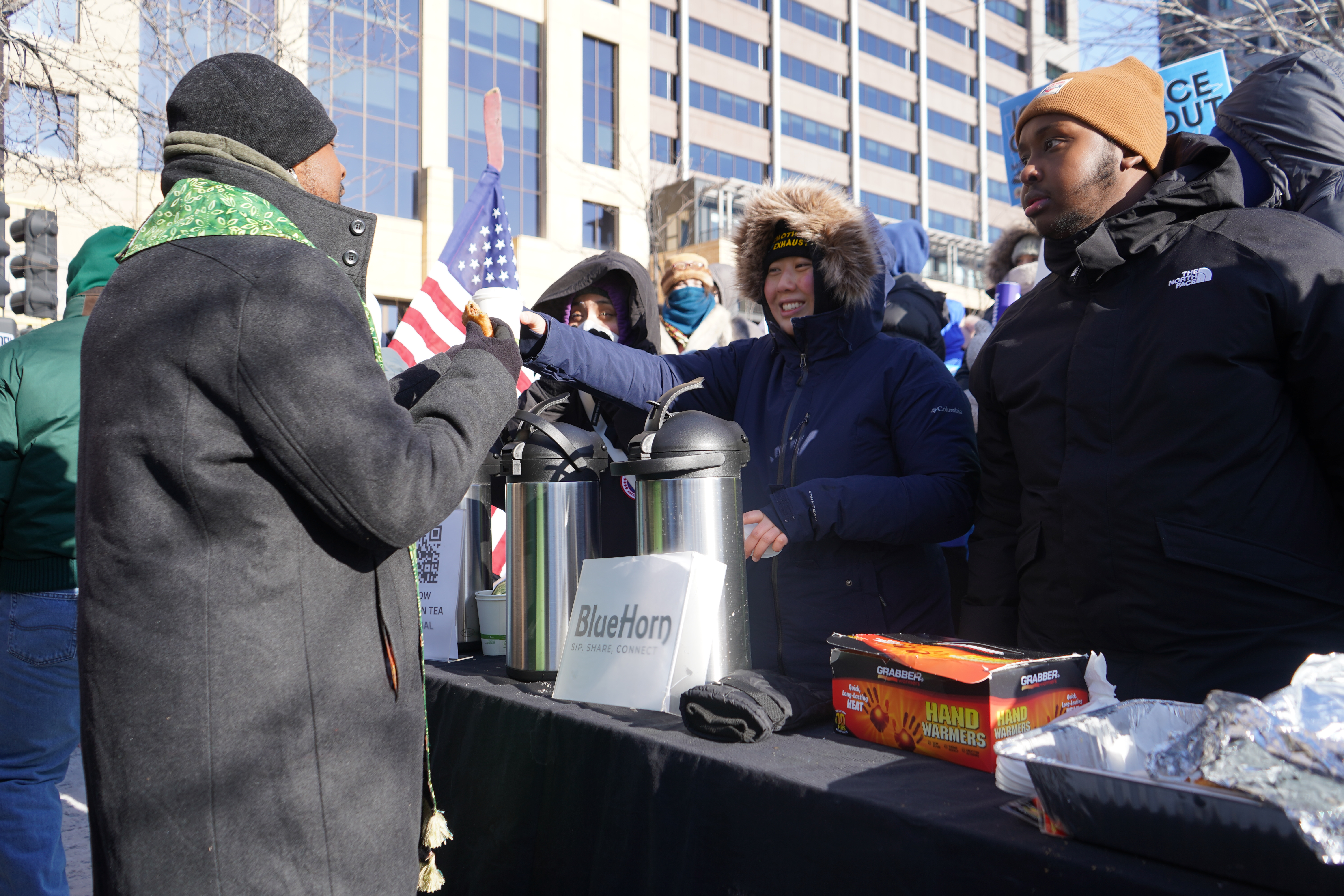 A volunteer hands a hot beverage to protestors, all clad in winter clothing.
