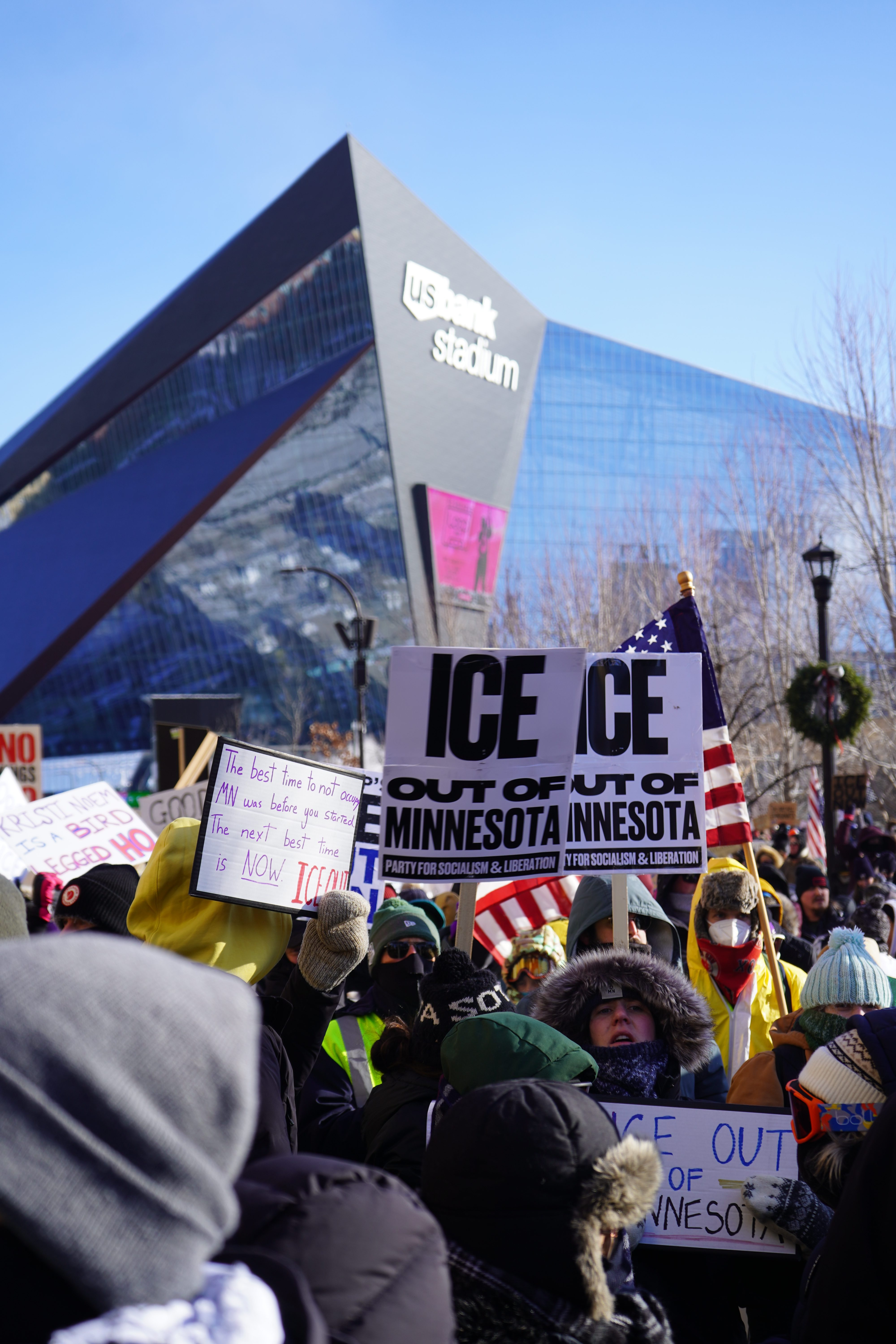 Protestors in winter coats and hats hold anti-ICE signage outside a stadium. 