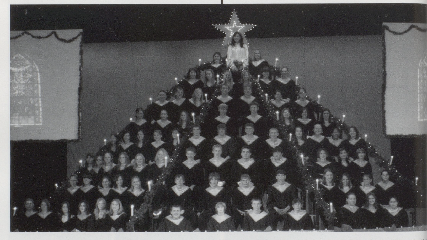 Yearbook photo of a choir group on bleachers, arranged in a pyramid to resemble a Christmas tree, with one female student dressed in white as the star at the top. A lit-up star sign hangs above her head.