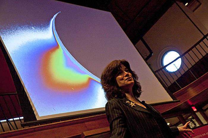 Carolyn Porco lecturing with an image of Saturn's moon Enceladus, giving off a plume, in the background.